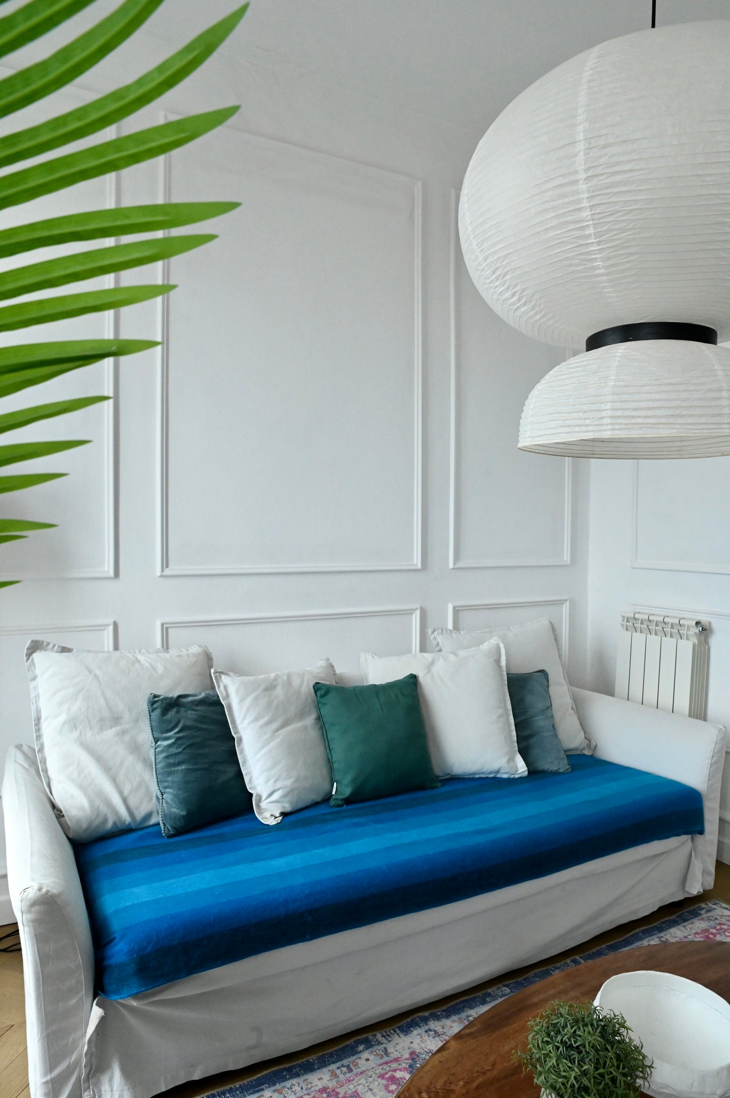 White sofa with blue alpaca throw blanket and green pillows in a room with white paneled walls and a hanging lamp.