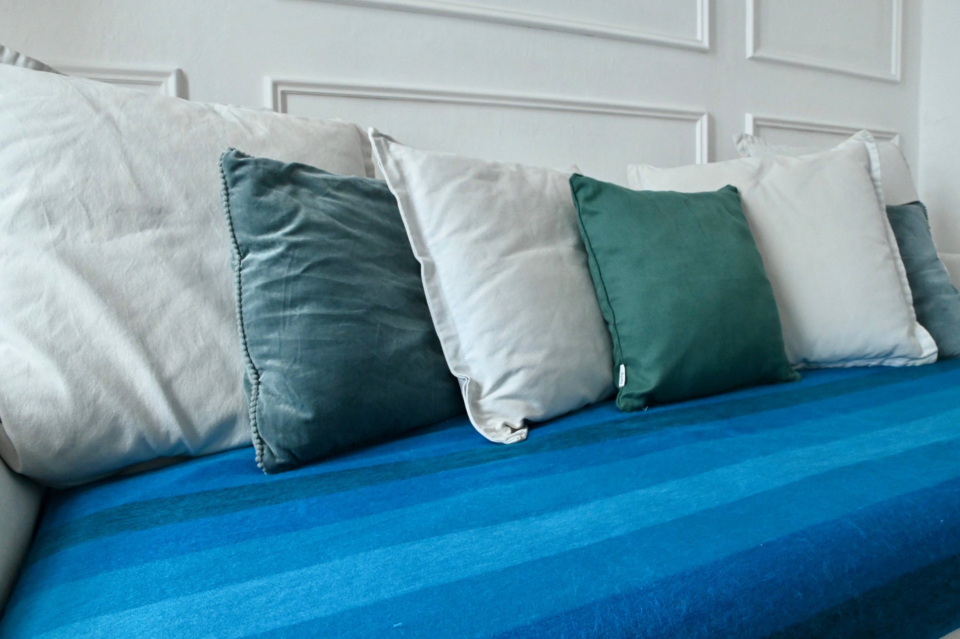 Blue striped alpaca blanket with green and white pillows on a sofa against a white paneled wall.