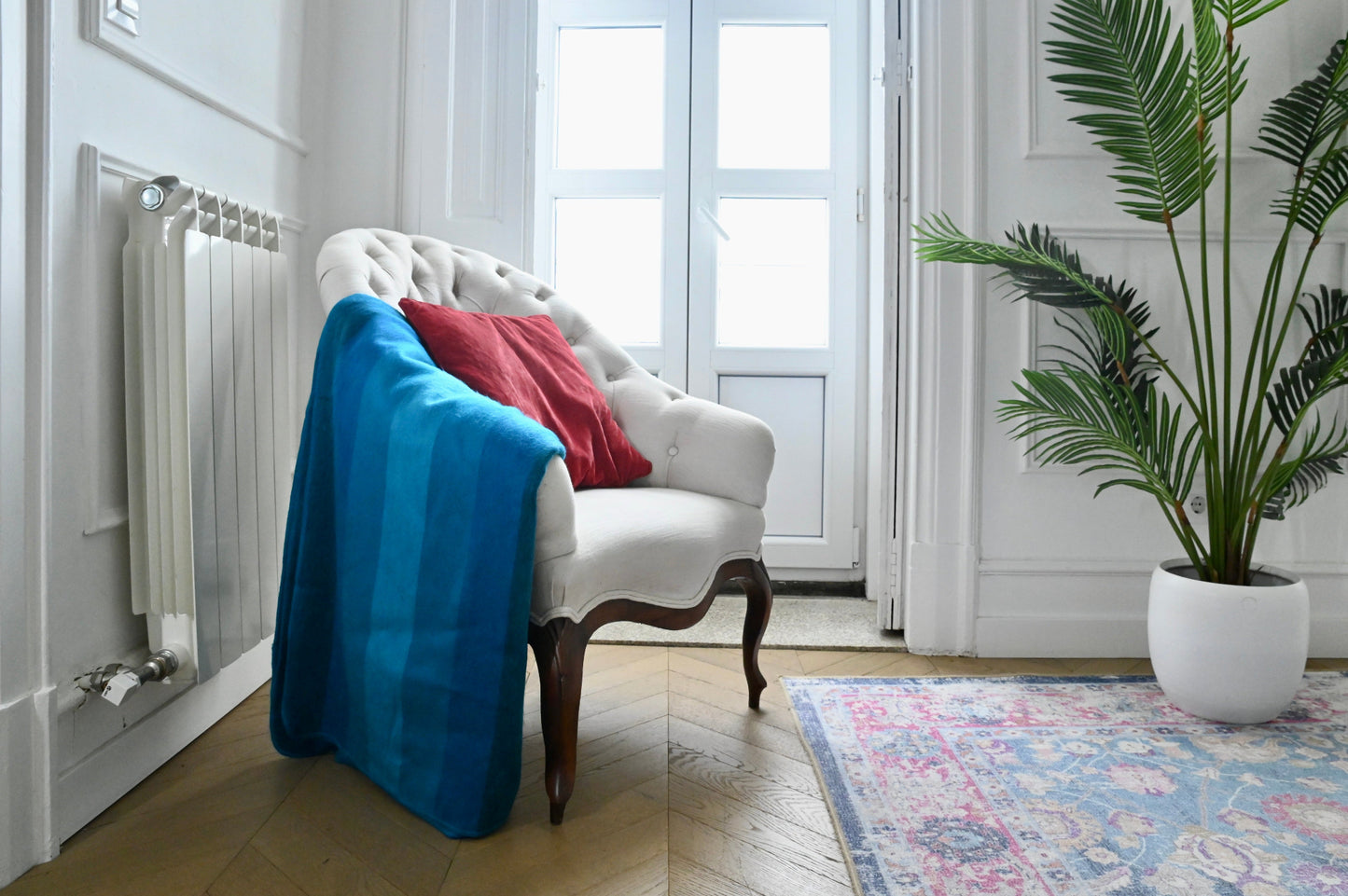 White armchair with a blue alpaca blanket and red pillow in a room with a plant and rug.