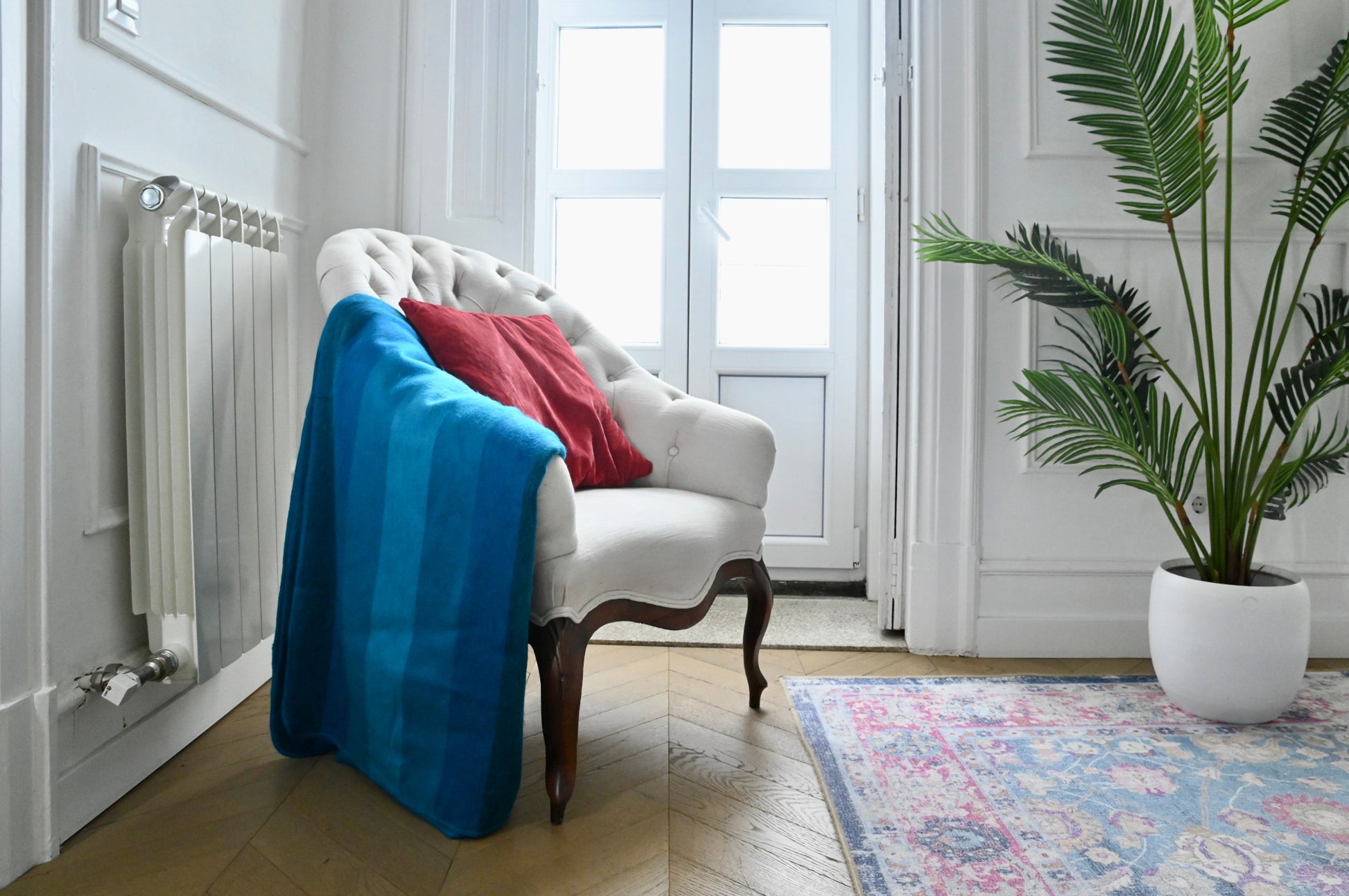 White armchair with a blue alpaca blanket and red pillow in a room with a plant and rug.