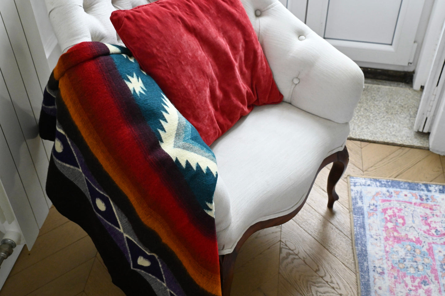 White armchair with a colorful alpaca blanket and red pillow on a wooden floor.