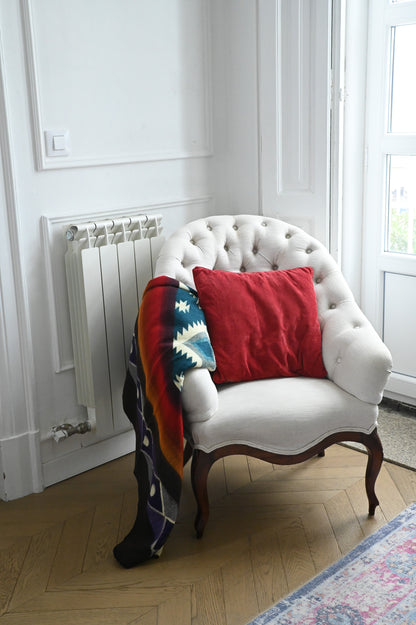 White chair with colorful alpaca blanket and red pillow against a window with white frame.