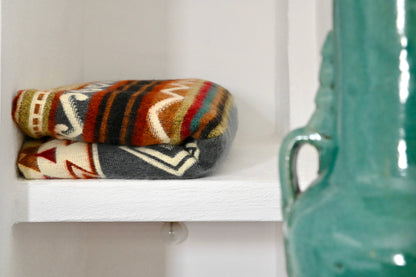 Colorful patterned alpaca blanket on a white shelf next to a teal ceramic vase.