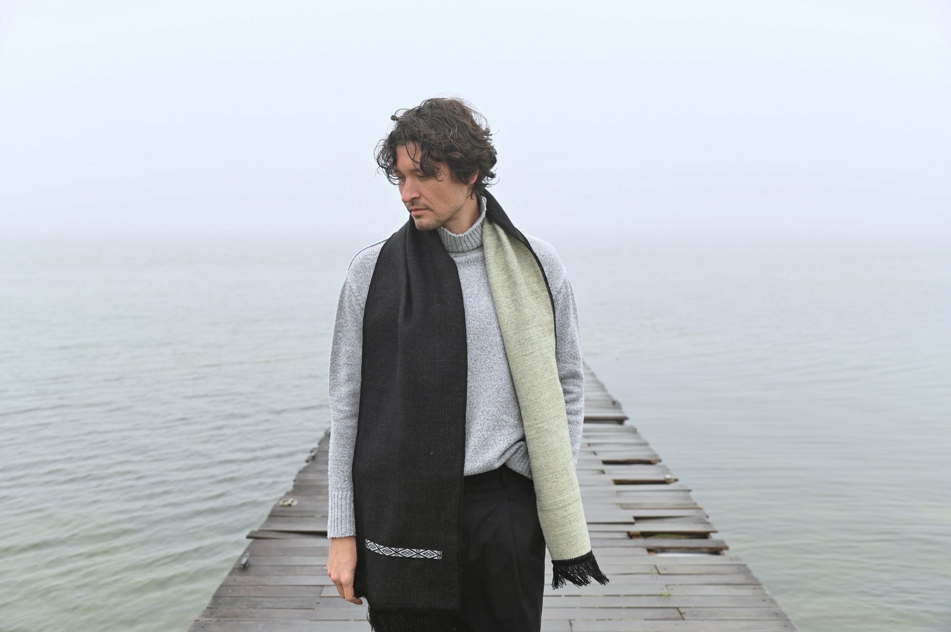 Person wearing a black and beige scarf on a wooden dock by a body of water