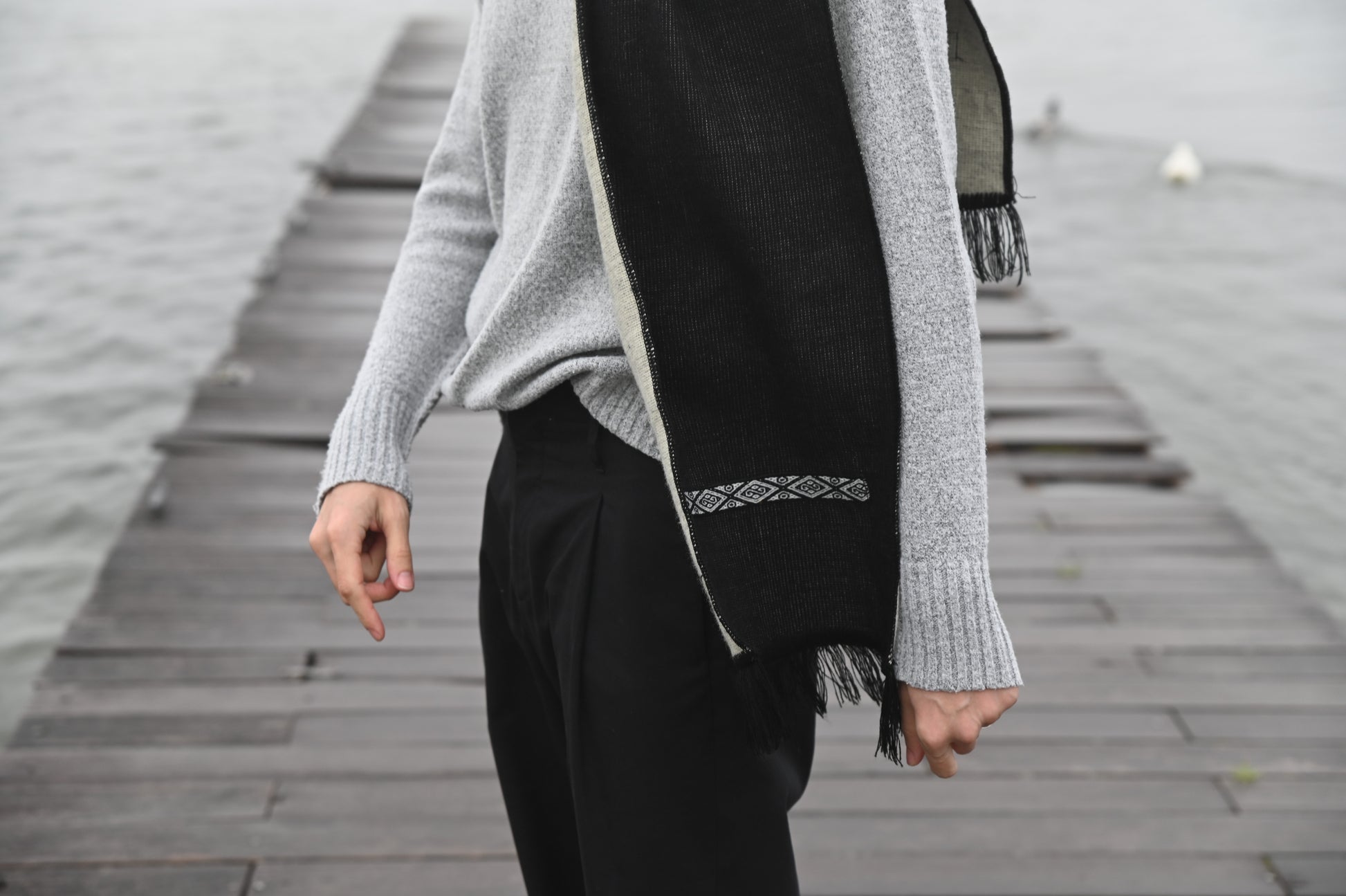 Person wearing a gray sweater and black scarf on a wooden dock by water